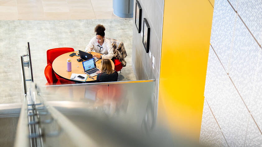 Two individuals collaborate at a round table in a modern, brightly lit space viewed from an elevated staircase.